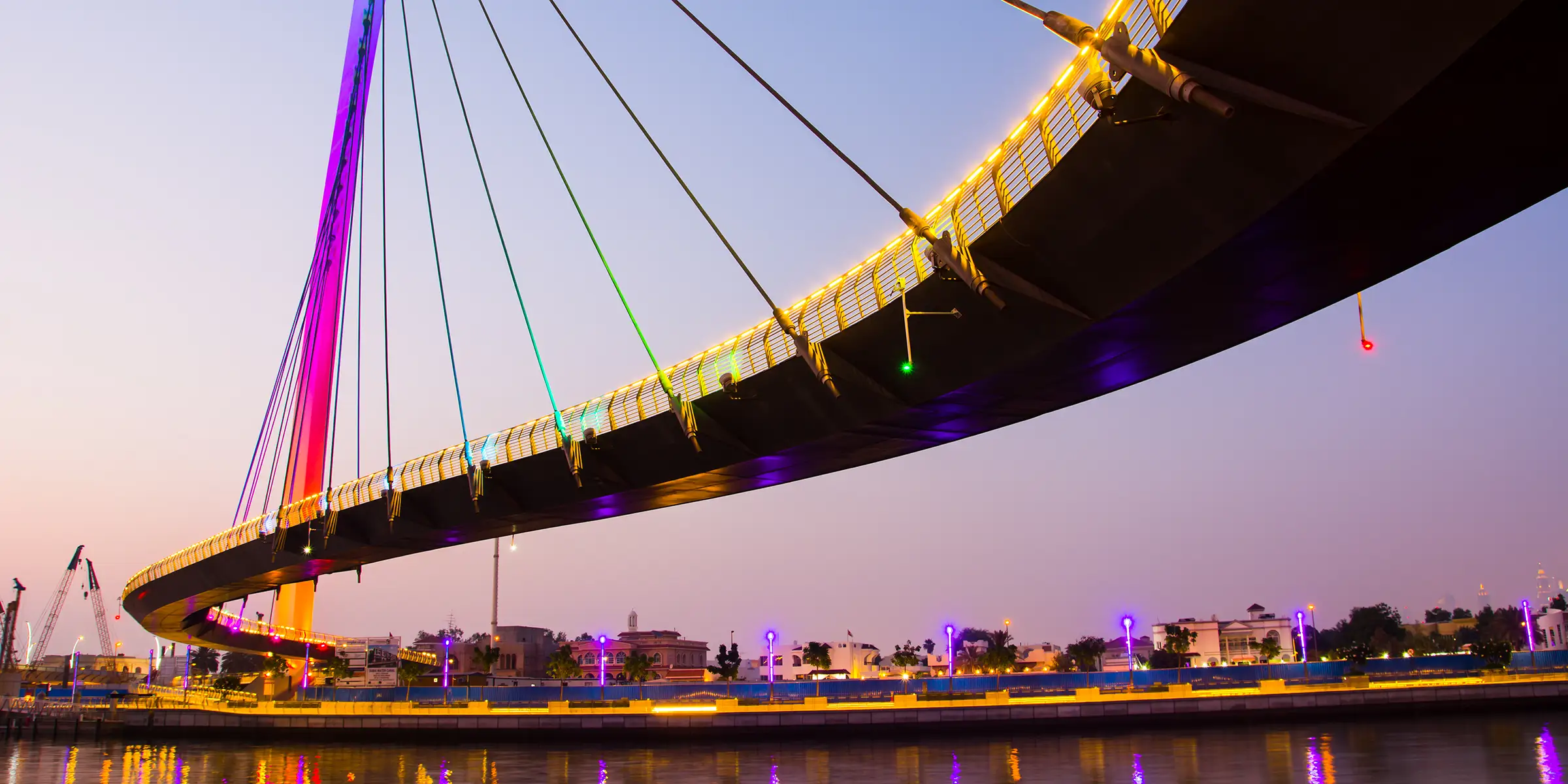 An illuminated cable-stayed bridge seen from below at dusk with city buildings beyond.