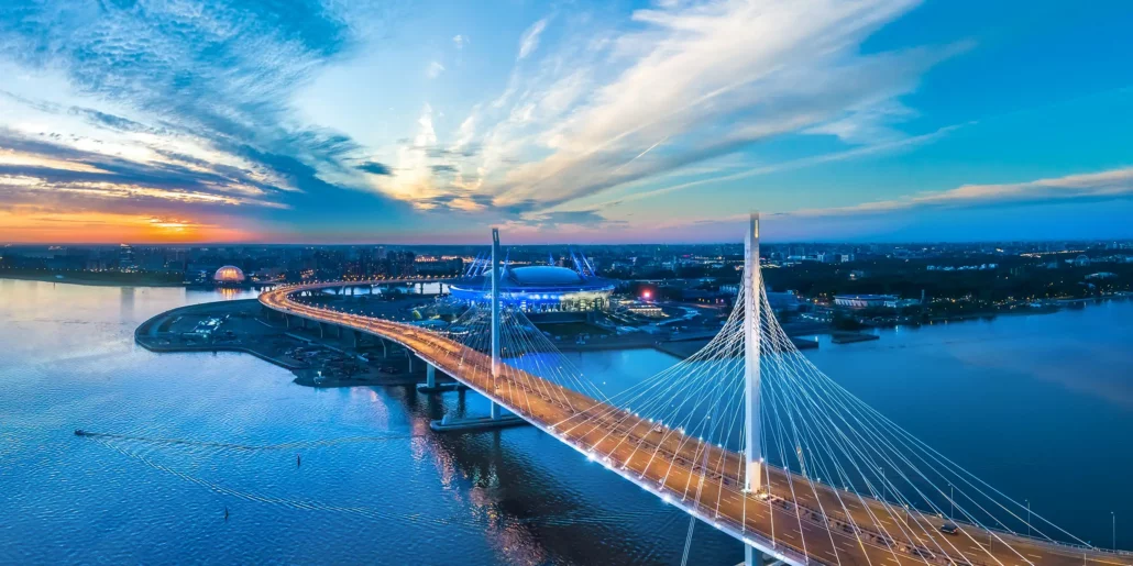 A cable-stayed bridge crossing a river at sunset with buildings and a stadium nearby.