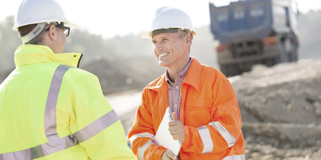 Two hard-hatted construction professionals in high-visibility jackets talking beside a truck at an earthworks site.