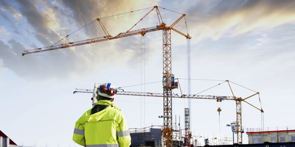 Construction professional in a high-visibility jacket looking towards tower cranes beneath a cloudy sky.