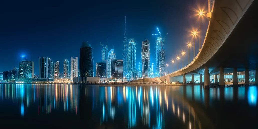Illuminated waterfront towers and a bridge reflected in calm water at night.