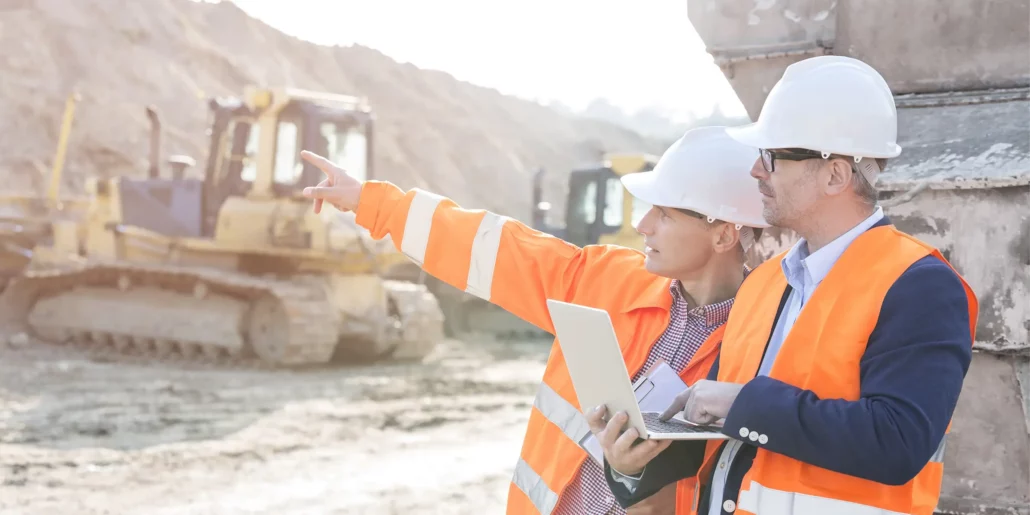 Two hard-hatted construction professionals reviewing a tablet as one points towards earthmoving equipment.